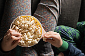 From above cropped unrecognizable young siblings kids hands sitting comfortable on sofa with popcorn bucket while eating and watching TV