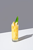 Glass full of cold pineapple juice with green leaves and straw placed on sunlit table against gray background