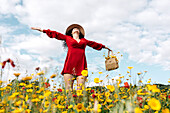 From below happy female in red sundress, hat and handbag standing with eyes closed on blossoming field with yellow and red flowers with outstretched arms enjoying on warm spring summer day