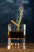 Ein Glas Whiskey mit Rosmarin auf einem Holztisch vor blauem Hintergrund