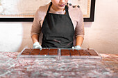 Crop woman in apron and gloves putting plastic molds with chocolate bars on table while working in professional confectionery