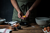 Unrecognizable guy in apron breaking raw egg over bowl while preparing pastry on lumber table near kitchenware