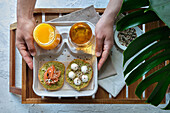 Top view of crop unrecognizable person eating appetizing healthy avocado toasts with salmon and burrata cheese served on tray with glasses of fresh juices in sunlight