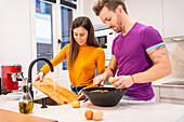 Woman washing wooden chopping board in sink while standing near boyfriend making salad in bowl while standing together in kitchen
