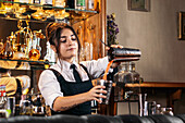 Focused female barkeeper pouring orange alcohol cocktail in metal shaker while preparing refreshing drink and standing at counter in bar