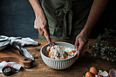 Unrecognizable male cook using whisk to mix pumpkin puree and cream with eggs and flour in bowl while preparing pie on timber table near cutting board and towel