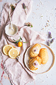Draufsicht auf leckere Madeleines auf einem Teller zwischen frischen Zitronenscheiben und blühenden Lavendelzweigen auf zerknittertem Stoff