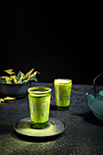 Stilleben mit traditionellem orientalischem Matcha-Tee, serviert in Gläsern mit Metalldekor auf einem Tisch mit Keramikschalen und frischen grünen Blättern vor schwarzem Hintergrund