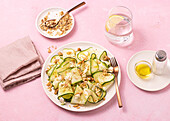 From above fresh salad of thin sliced ripe zucchini, feta cheese and pine nuts in white plate on table pink background