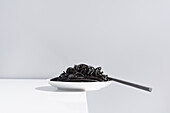 Stainless fork in full bowl of black spaghetti with cuttlefish ink on white table in studio on gray background