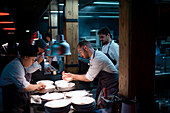 Young cooks men in aprons carefully serving meal in white ceramic dishes on restaurant kitchen
