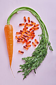 From above composition of scattered vitamin pills arranged on pink table near ripe carrots