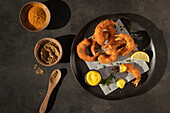 From above still life with plate of lemon and mustard battered prawns with two bowls of turmeric and curry spices