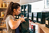 Side view of female seller in apron with ponytail standing near shelves with containers while checking quality of food during working day