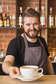 Bearded male barista in apron serving cup of coffee while working at bar table in coffee shop
