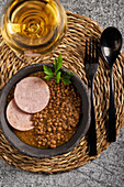 Rustikale Schale mit leckerer Linsensuppe mit Petersilie und Wurstscheiben auf Marmortisch neben Wein und Besteck beim Mittagessen