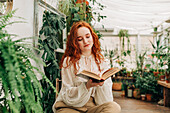 Concentrated red haired female sitting on chair with crossed legs and reading book among different potted plants in domestic garden