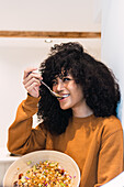 Positive young African American female with curly dark hair in sweater eating tasty vegetable salad in kitchen at home and looking away
