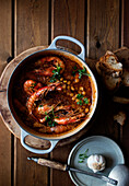 Top view of white saucepan with delicious homemade stew with chickpeas and shrimps placed on round wooden tray next to rustic metal plate with ladle and garlic and some croutons on wooden background