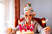 Positive aged female in sunglasses and with glowing Bengal lights sitting at table with cake with 100 candle and celebrating anniversary while looking at camera