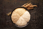 From above of bowl with assorted types of dried rice placed near bunch of ears and sickle on black background