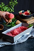 Anonymous person showing wafer cone with ball of watermelon sorbet near green plant over table with container and napkin