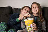 Young cheerful siblings kids sitting comfortable on sofa with popcorn bucket while eating and watching TV