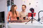 Positive couple of bloggers recording vlog on smartphone with microphone while sitting at kitchen counter with cup of coffee at home