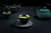 Schwarze Keramiktasse mit traditionellem japanischen grün gefärbten Matcha-Tee, serviert auf einem Tisch mit Teekanne