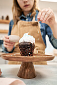 Crop anonymous female decorating tasty chocolate cupcake on cake stand with sugar sprinkles while cooking in kitchen