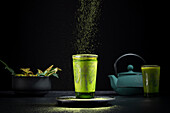 Stillleben Komposition mit traditionellem orientalischen Matcha-Tee in Glastasse mit Metallverzierung auf Tisch mit Keramikschalen und frischen grünen Blättern vor schwarzem Hintergrund