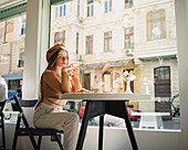 French female in beret sitting at table in cafe with aromatic glass of coffee and freshly baked croissant