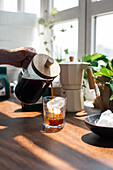 Crop unrecognizable person pouring freshly brewed coffee from glass coffee press into glass with ice cube on wooden table in light kitchen