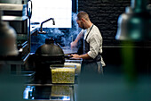 Junger Mann in grauer Schürze rührt in einer großen Metallpfanne in einer Restaurantküche