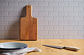 Wooden chopping board placed near wall on table with bowl and knife in light kitchen