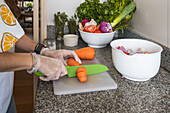 Crop anonymous female in gloves cutting slices of ripe orange carrot on chopping board with salad while preparing healthy vegetarian dish on counter with assorted vegetables in kitchen