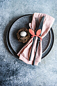 Table set for Easter dinner with pink napkin and a bird nest with an egg on a concrete background
