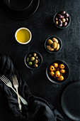 From above tasty juicy green brown yellow olives and oil in black and white bowls on black table decorated with tablecloth and forks