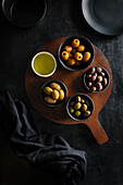 From above juicy green brown yellow olives and oil on round wooden stand on black table decorated with tablecloth