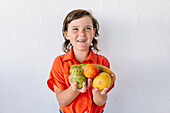 Adorable little girl in bright red shirt happily looking away while demonstrating assorted fresh exotic fruits against white background