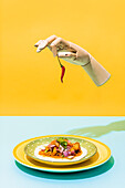 Artificial hand with red hot chili pepper levitating over plate with Mexican chicken salad on blue and yellow background