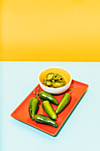 From above raw and marinated jalapenos placed on red plate on yellow and blue background