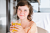 Braunhaariges Mädchen im Vorschulalter schaut in die Kamera und trinkt leckeren Orangensaft aus einem Glas in einer Cafeteria