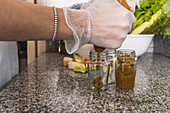 Side view of crop anonymous female in gloves pouring vegetable appetizer in glass bottle near bowl with green lettuce in kitchen at home