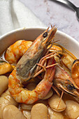 Schale mit typischem Bohneneintopf mit Garnelen, Krabben und Muscheln auf einer Spitzentischdecke neben einem Löffel und auf einem Steintisch