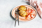 From above baked Hasselback spiced potatoes with bacon slices served in pink ceramic bowl on concrete background