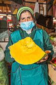 Ältere peruanische Verkäuferin mit medizinischer Maske blickt in die Kamera und zeigt einen aufgeschnittenen reifen gelben Kürbis neben verschiedenen Produkten, während sie auf einem Wandermarkt arbeitet