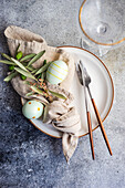 From above cutlery set for Easter dinner with olive tree branches and eggs on plate on a concrete background