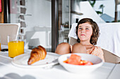 Wistful child with brown hair sitting at table with plates of delicious food in cafeteria and looking at camera
