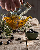 Unbekannter Koch gießt frisches Öl auf reife schwarze Oliven über Holztisch in der Nähe von Zweigen